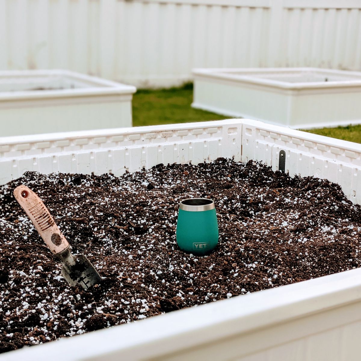 Giantex White raised garden bed with soil, hand spade, and wine glass with fence and more garden boxes in the distance